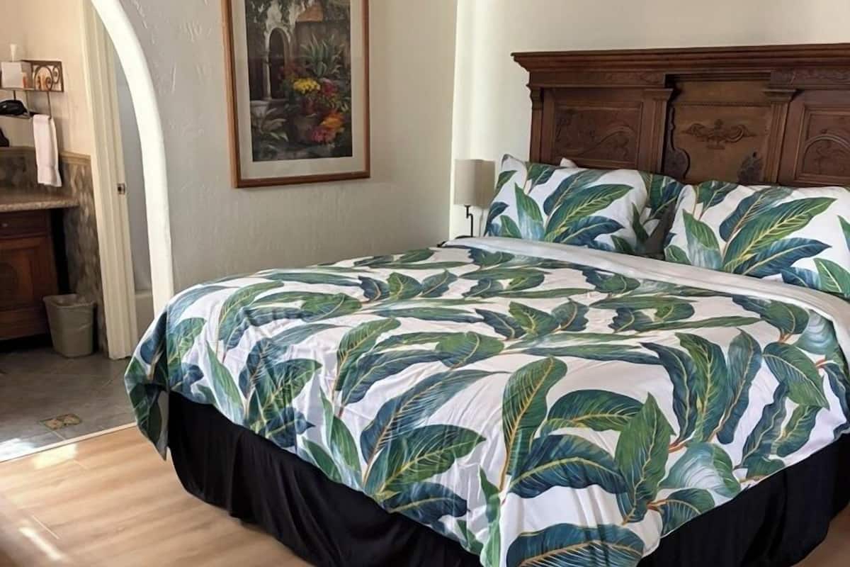 A bedroom with a bed featuring a tropical leaf pattern bedspread, a wooden headboard, and a framed painting on the wall.