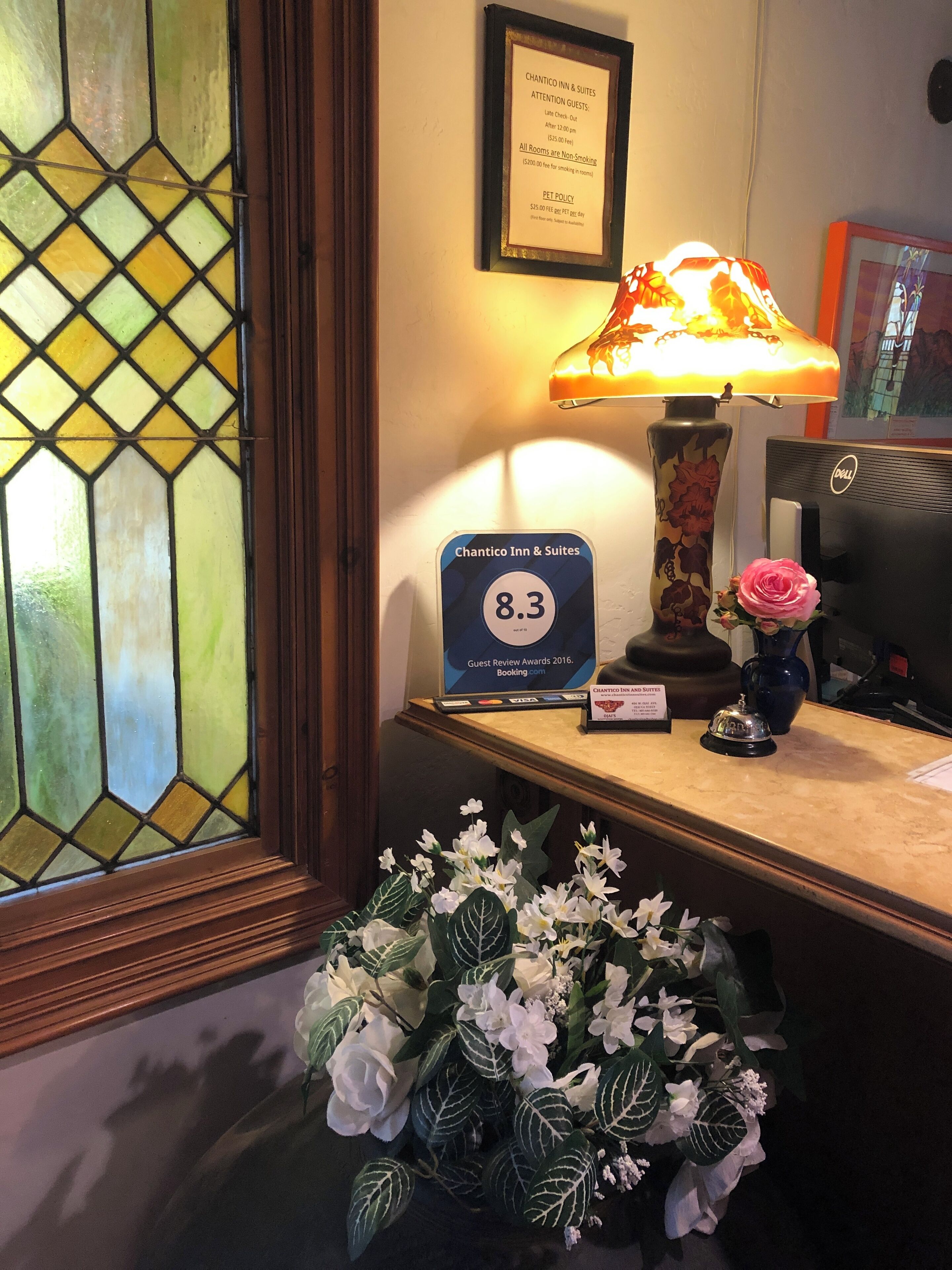 A reception desk with a framed guest review award, a lamp, a Dell computer, and a floral arrangement.