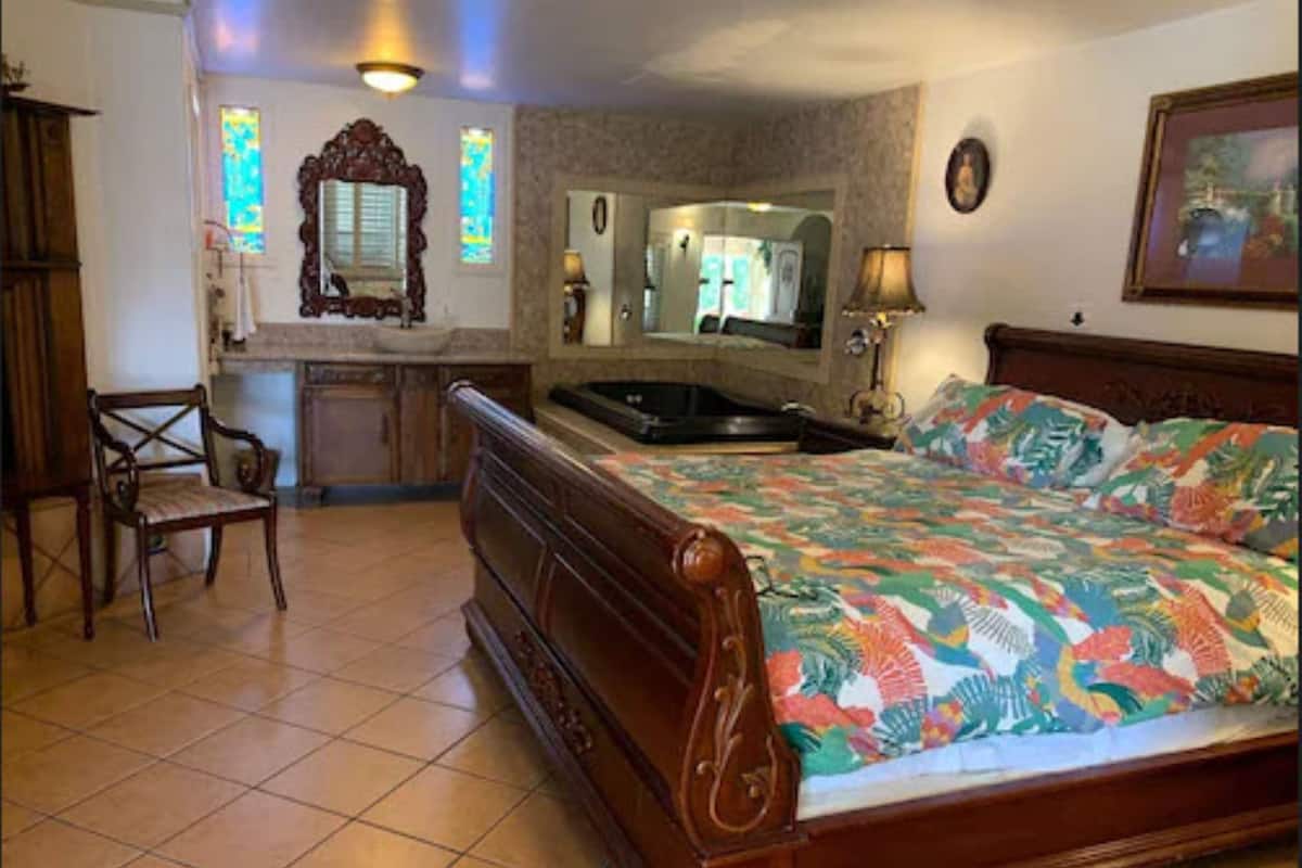 A bedroom with a wooden bed, a chair, a small desk, a mirror, and a window with stained glass.
