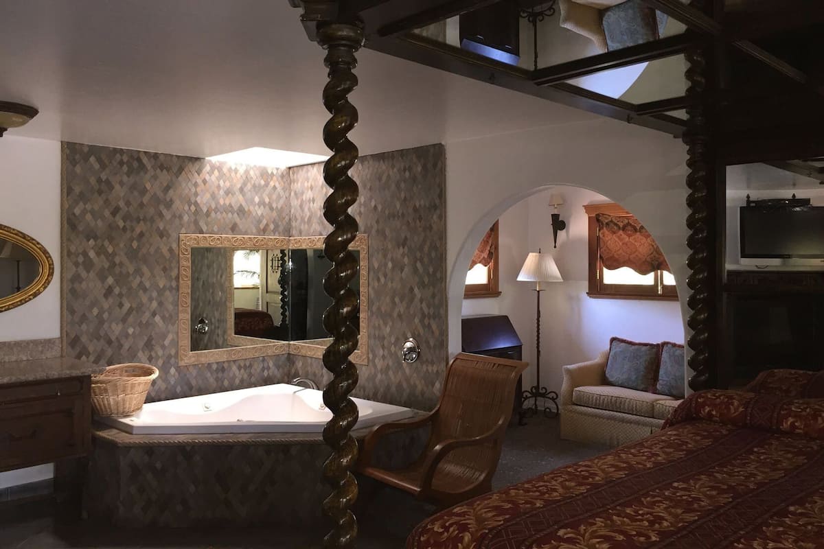 A bedroom with a large bed, a sitting area with a chair and a small table, a bathtub, a mirror, and a television.
