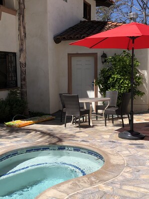 A swimming pool with a tiled edge, surrounded by stone flooring, and a dining area with wicker chairs and a table under a red umbrella.
