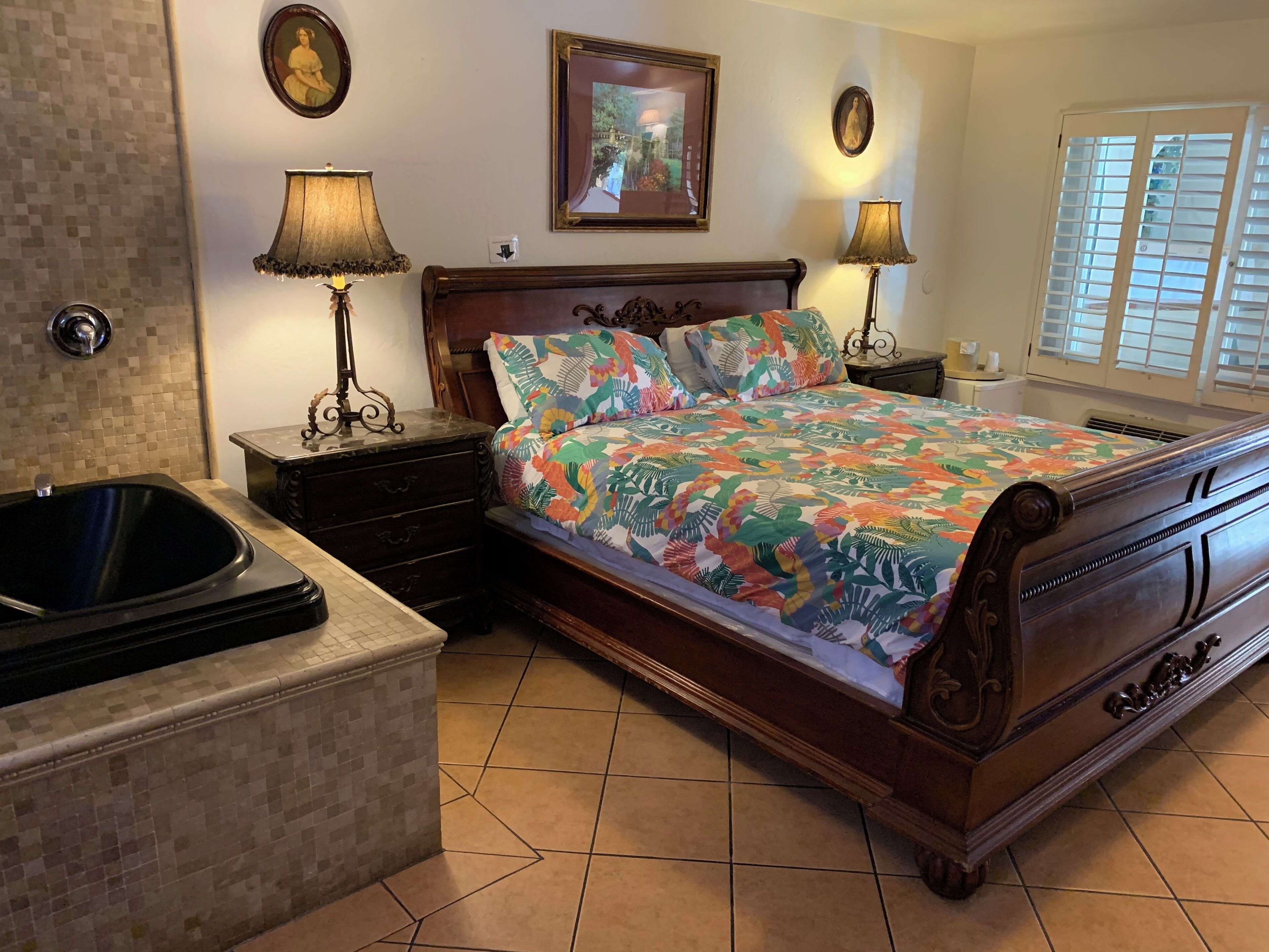A bedroom with a wooden bed, two bedside lamps, a nightstand, a decorative mirror, and a window with blinds.