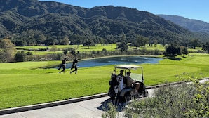 A golf course with a lake, mountains in the background, and a golf cart on the road.