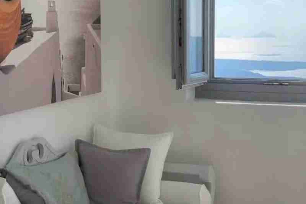 A room with a white sofa, a window with a view of the sea, and a wall-mounted shelf with a vase.