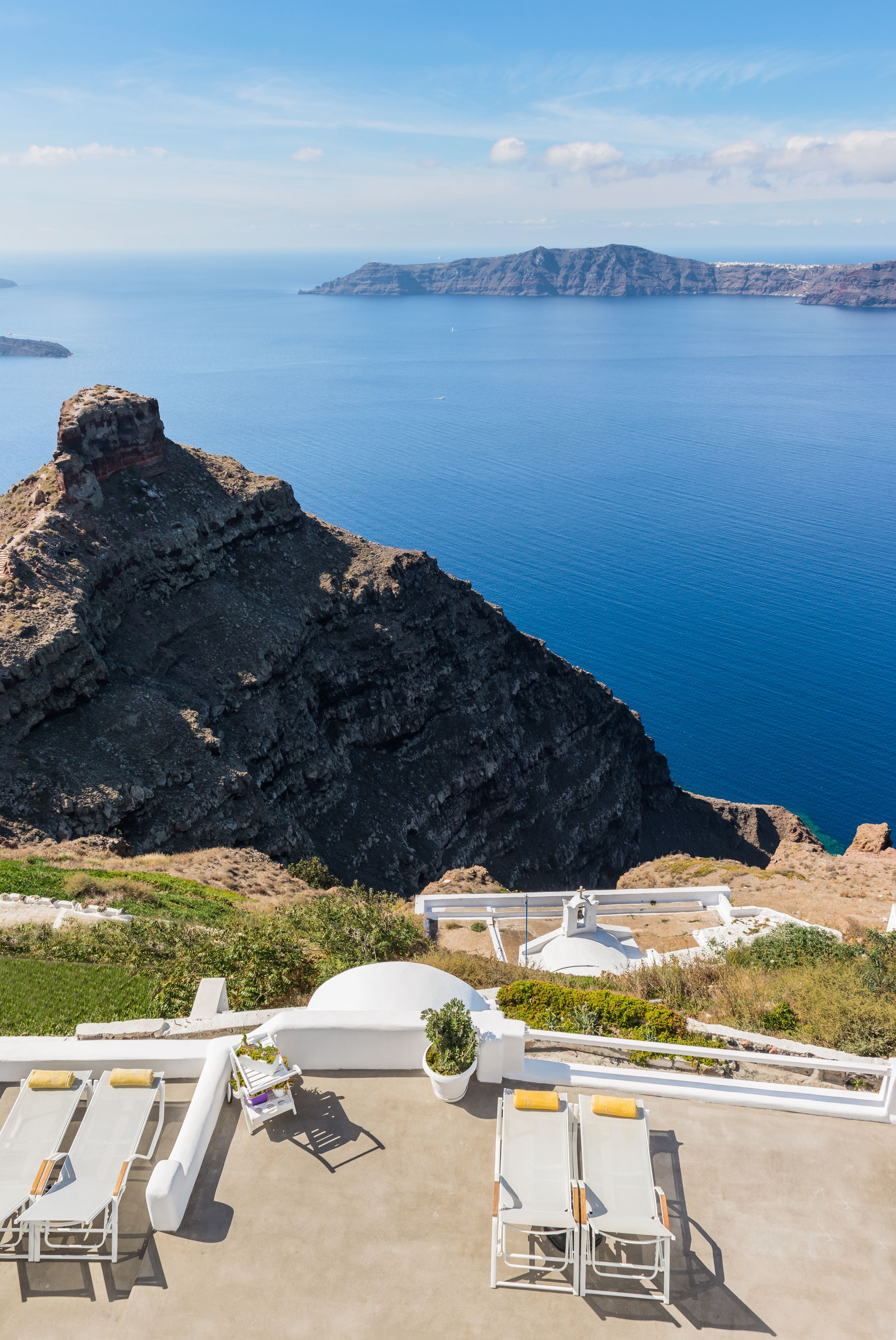 A seaside terrace with white lounge chairs and a white dome structure, overlooking a rocky coastline and the sea.