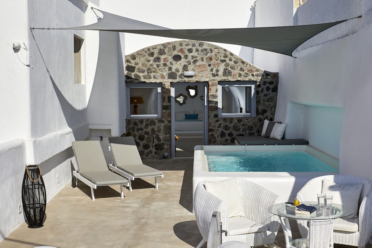 A sunlit outdoor area with a stone wall, a pool, lounge chairs, and a table with a book and glass.