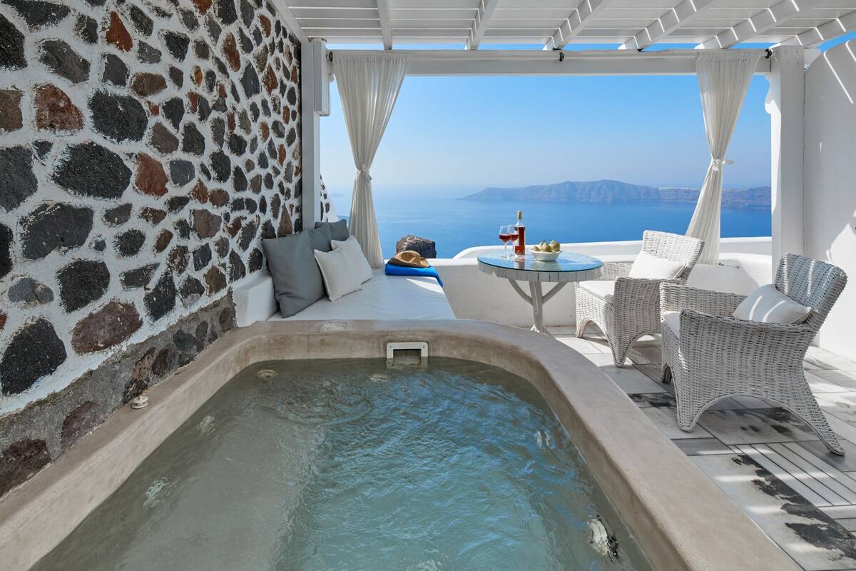 A stone-walled room with a hot tub, white wicker furniture, and a table with drinks.