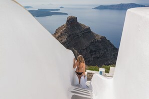 A person in a bikini standing on a white staircase with a view of a rocky coastline and the sea. - Iliovasilema Suites (Santorini)