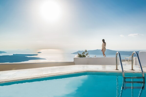 A pool with clear blue water, a person standing by the pool, and a view of the sea and mountains in the background. - Iliovasilema Suites (Santorini)