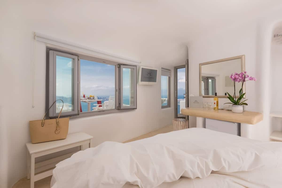 A bedroom with a bed, a bedside table, a vanity with a mirror, a window with a view, and a handbag on a bench.