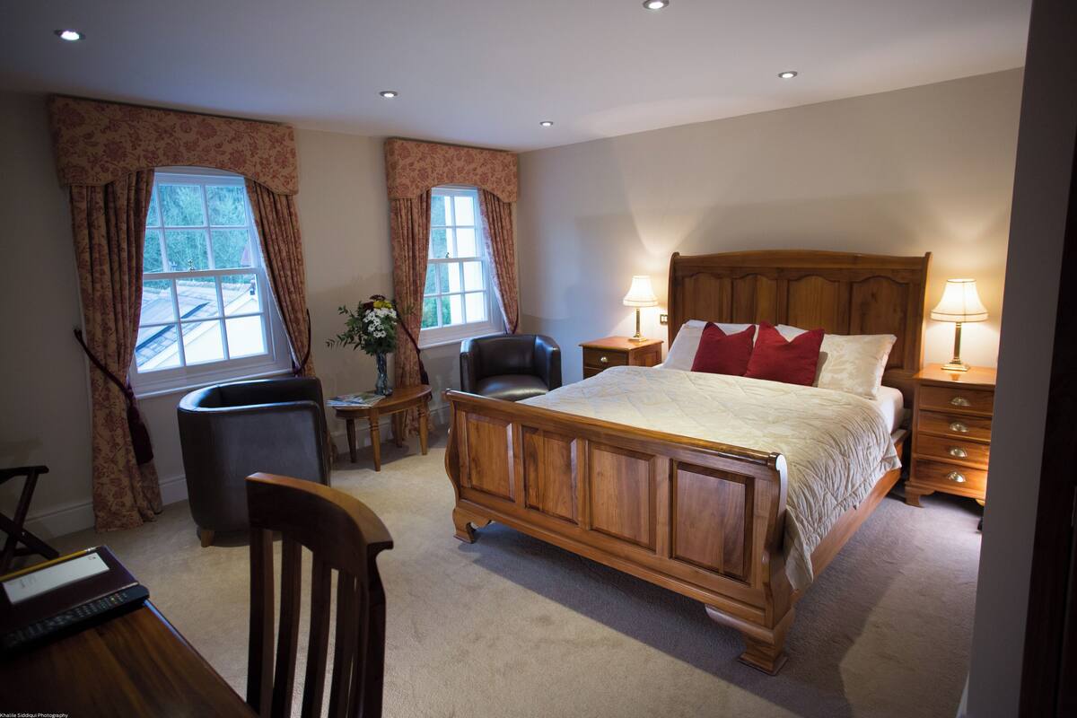 A bedroom with a wooden bed, two bedside tables with lamps, a chair, a desk with a computer, and a window with curtains.