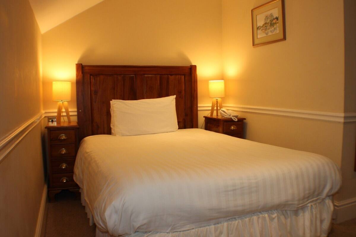 A neatly made bed with white linens in a hotel room. There are two bedside tables with lamps and a wooden headboard.