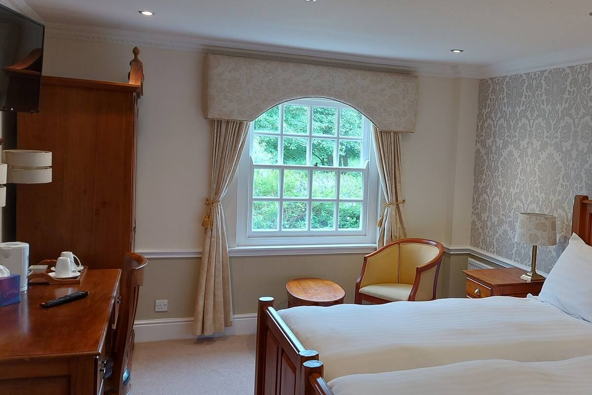 A hotel room with a large bed, a wooden nightstand, a chair, a desk with a lamp, and a window with curtains.
