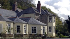 A large, multi-story building with a stone facade and multiple chimneys. There is a stone wall in front of the building, and a grassy area with outdoor furniture.