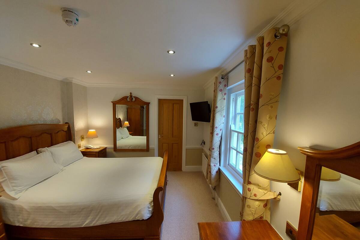 A hotel room with a large bed, a bedside table, a mirror, a television mounted on the wall, and a window with curtains.