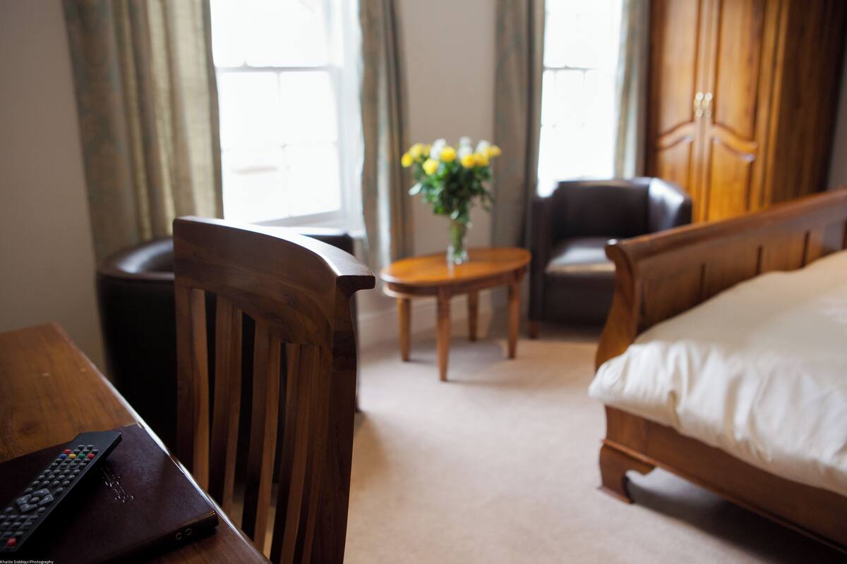 A hotel room with a wooden bed, a small round table with a vase of flowers, a chair, and a desk with a remote control.