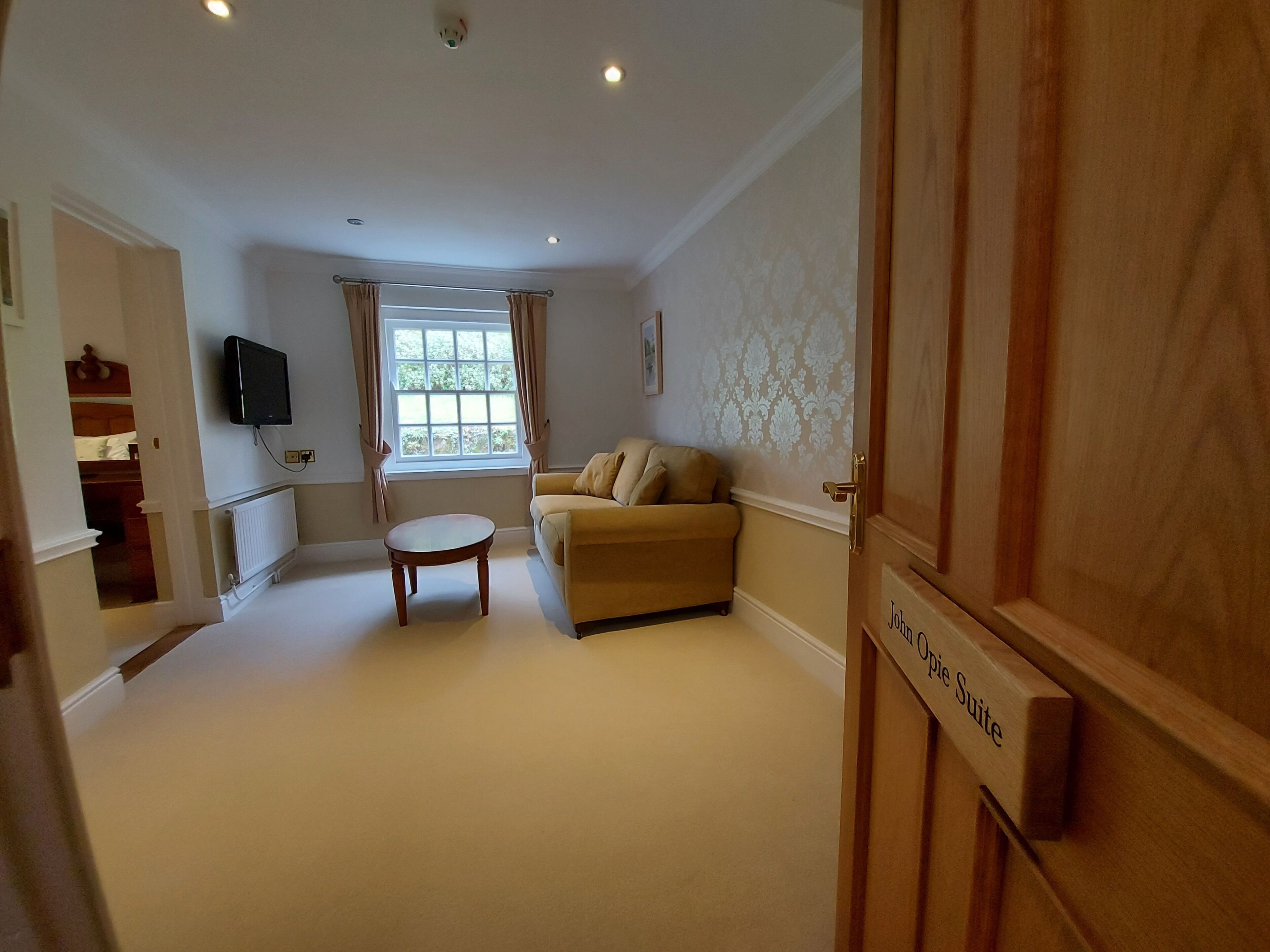 A living room with a beige sofa, a small round table, a window with curtains, a television mounted on the wall, and a door labeled "John Opie Suite".