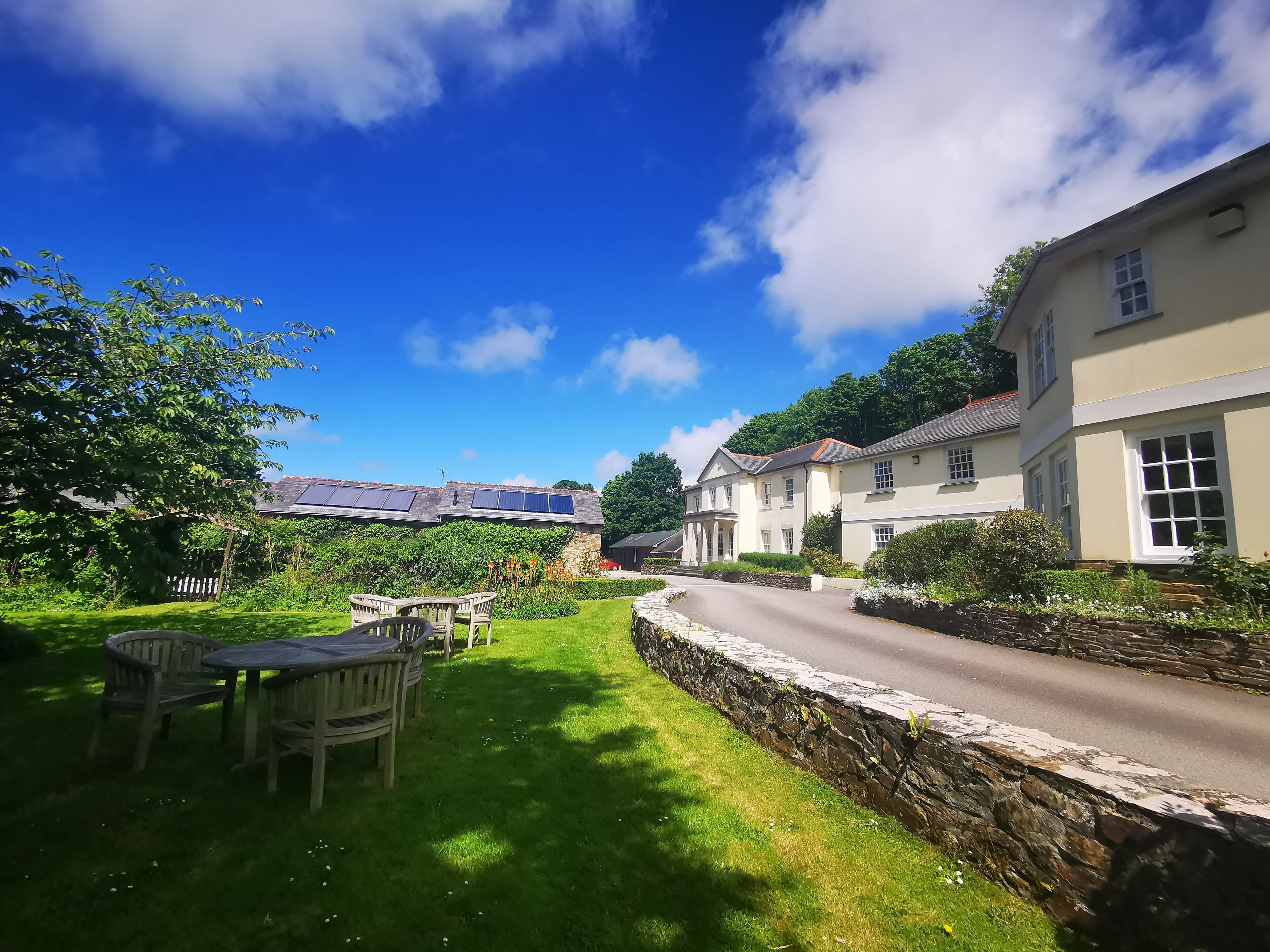 A traditional house with a stone wall, a paved driveway, and a grassy garden with a wooden table and chairs.