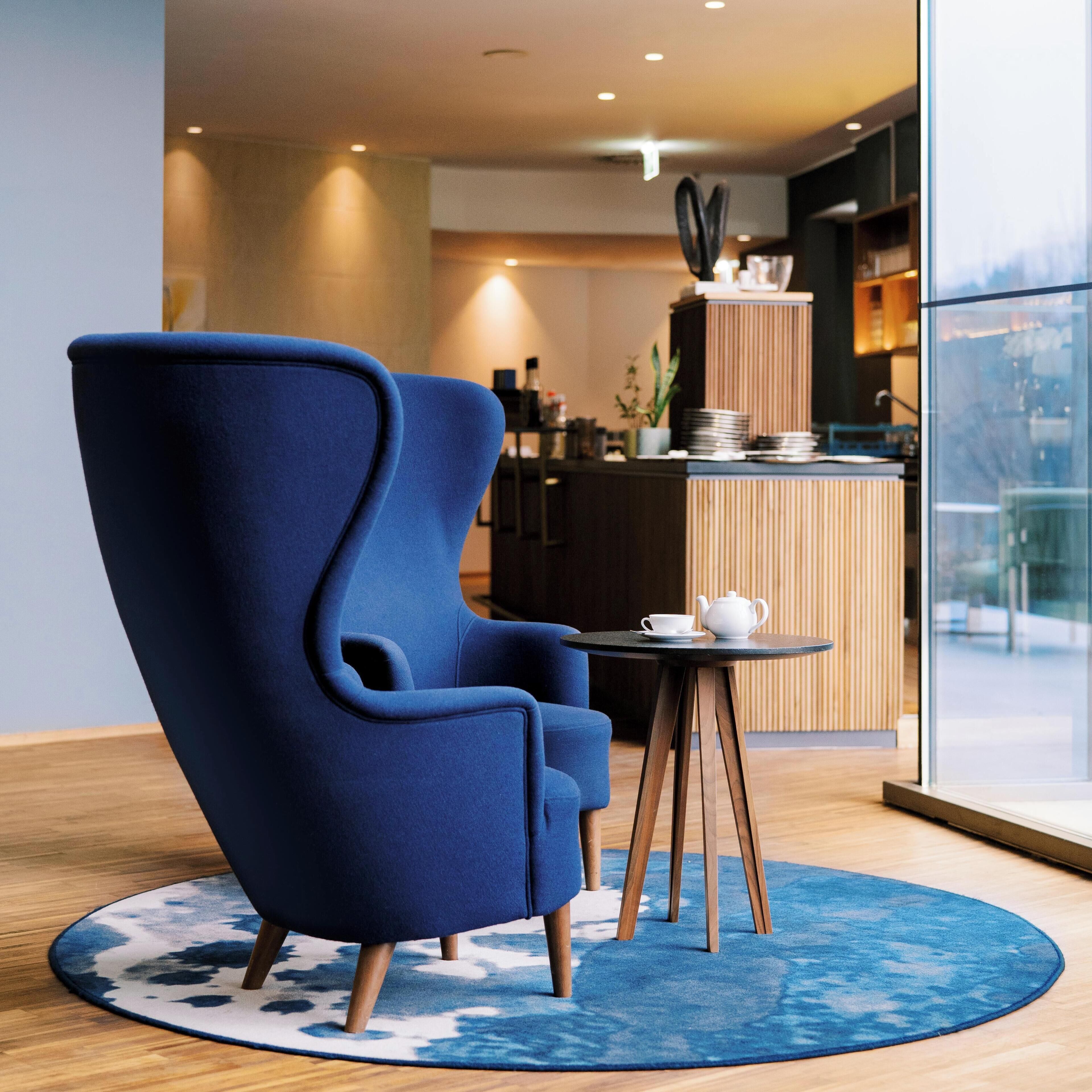 Una habitación moderna con un sillón azul, una mesita redonda con una tetera y vistas a la ciudad a través de amplios ventanales.
