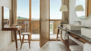 A modern bathroom with a wooden vanity, a large mirror, and a view of the outdoors through a sliding door. - Falkensteiner Balance Resort Stegersbach (Stegersbach)