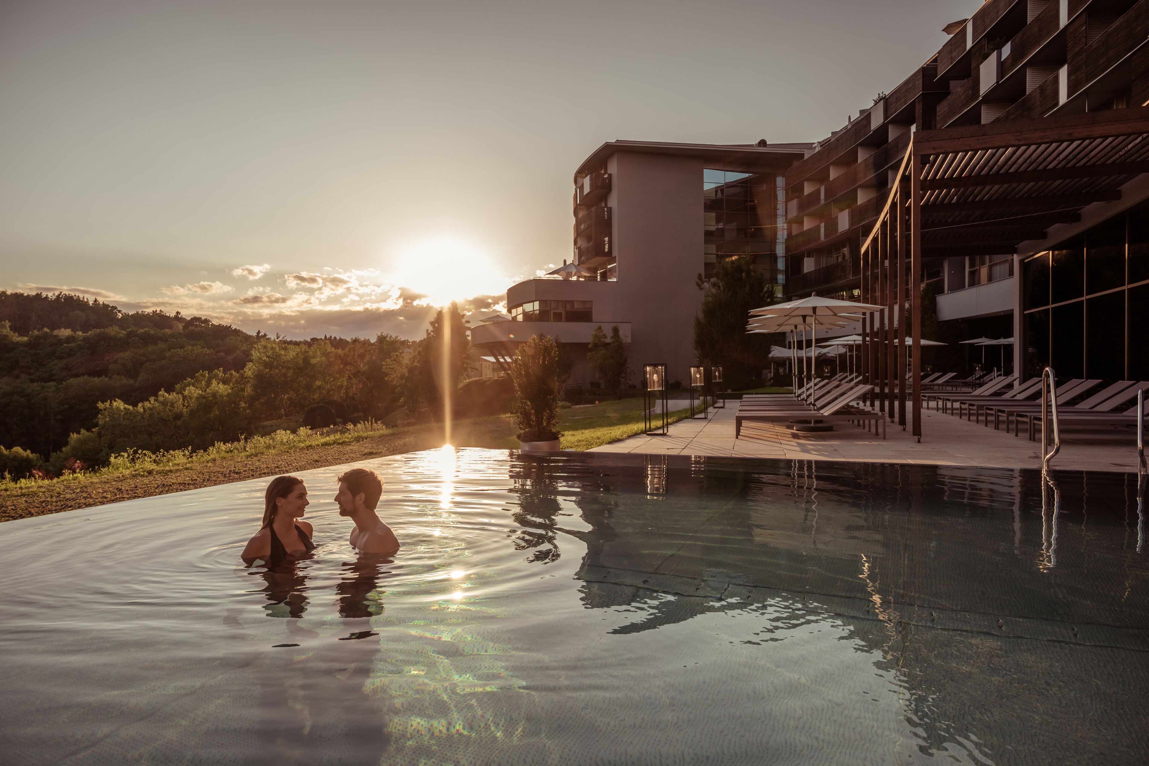 A swimming pool with a sunset in the background, surrounded by a modern building with balconies and outdoor seating.