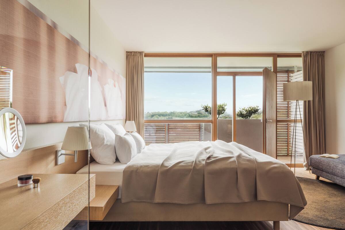 A modern hotel room with a large bed, a bedside table with a lamp, a view of the outdoors through a sliding door, and a seating area with a sofa.