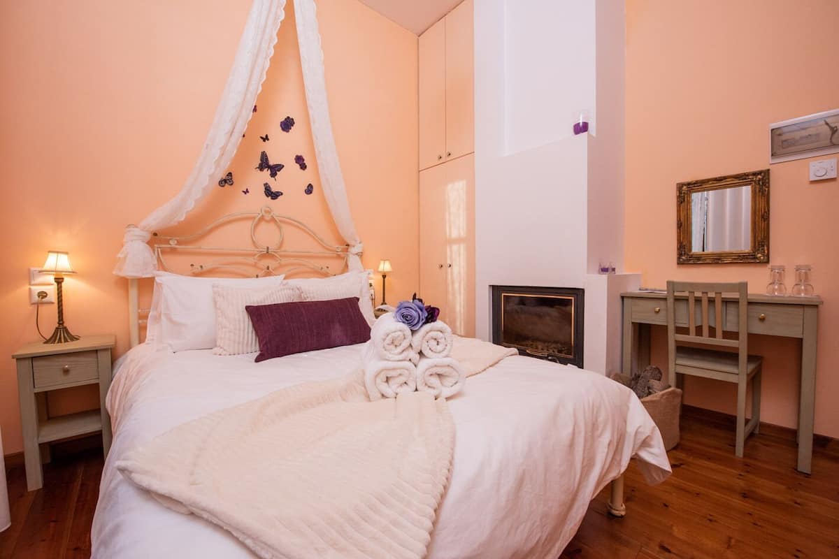 A bedroom with a canopy bed, a bedside table with a lamp, a desk with a chair, and a fireplace.