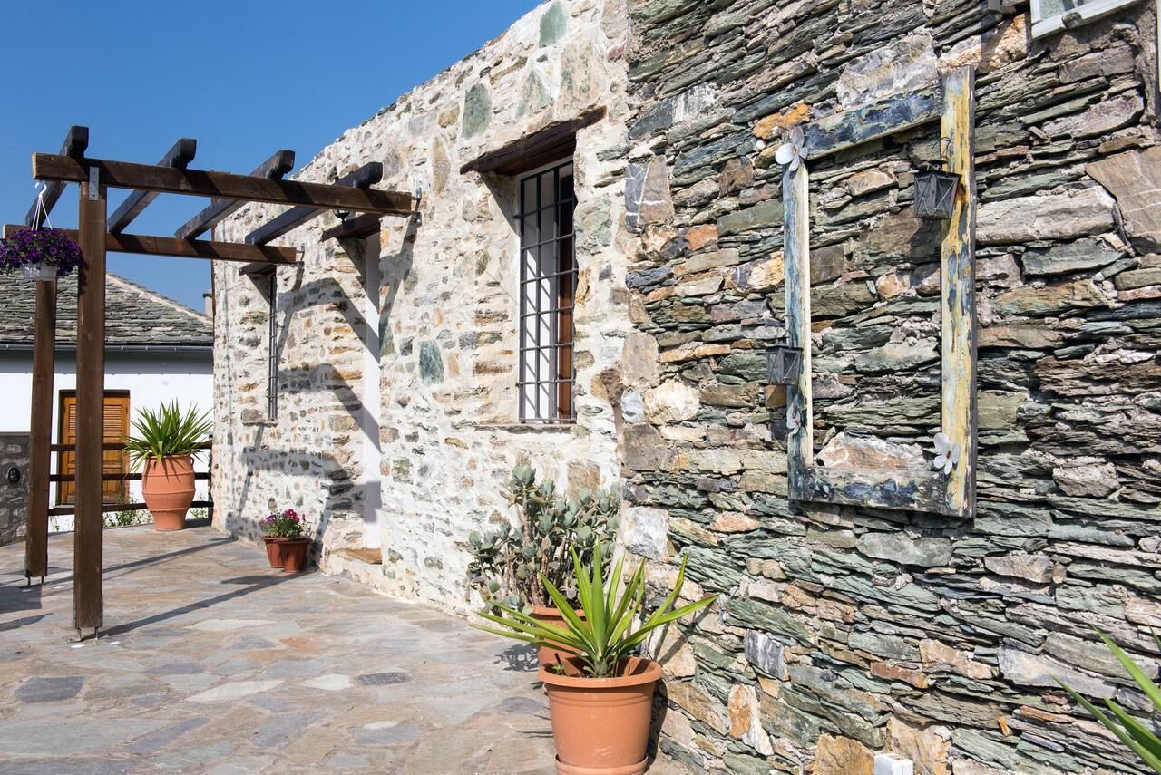 A stone building with a wooden pergola, potted plants, and a window with a metal grille.