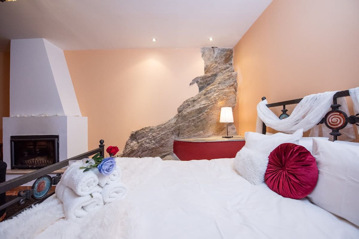 A bedroom with a large bed, a fireplace, a stone wall, and a nightstand with a lamp.