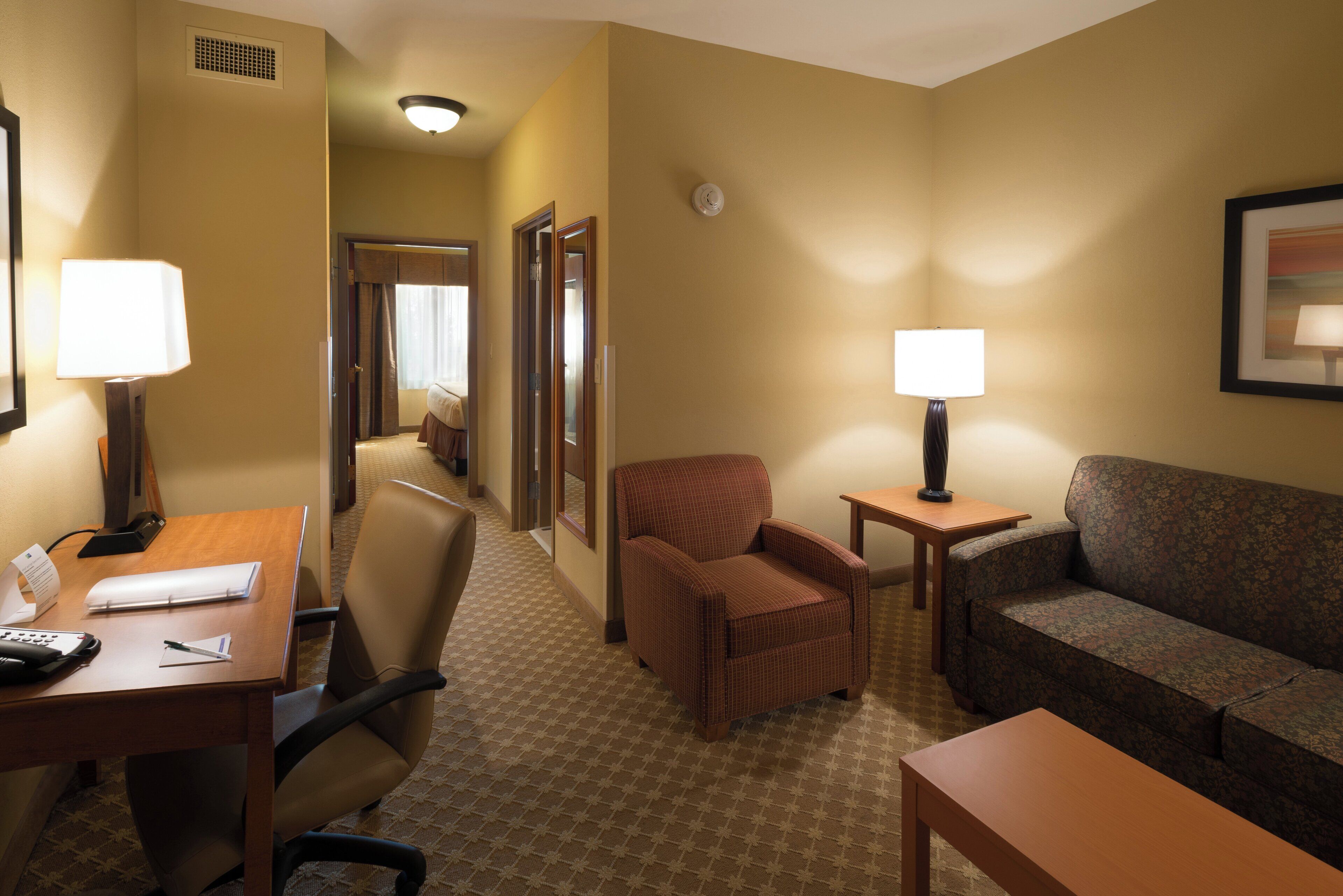 A hotel room with a desk, two chairs, a sofa, a small table, and a lamp.
