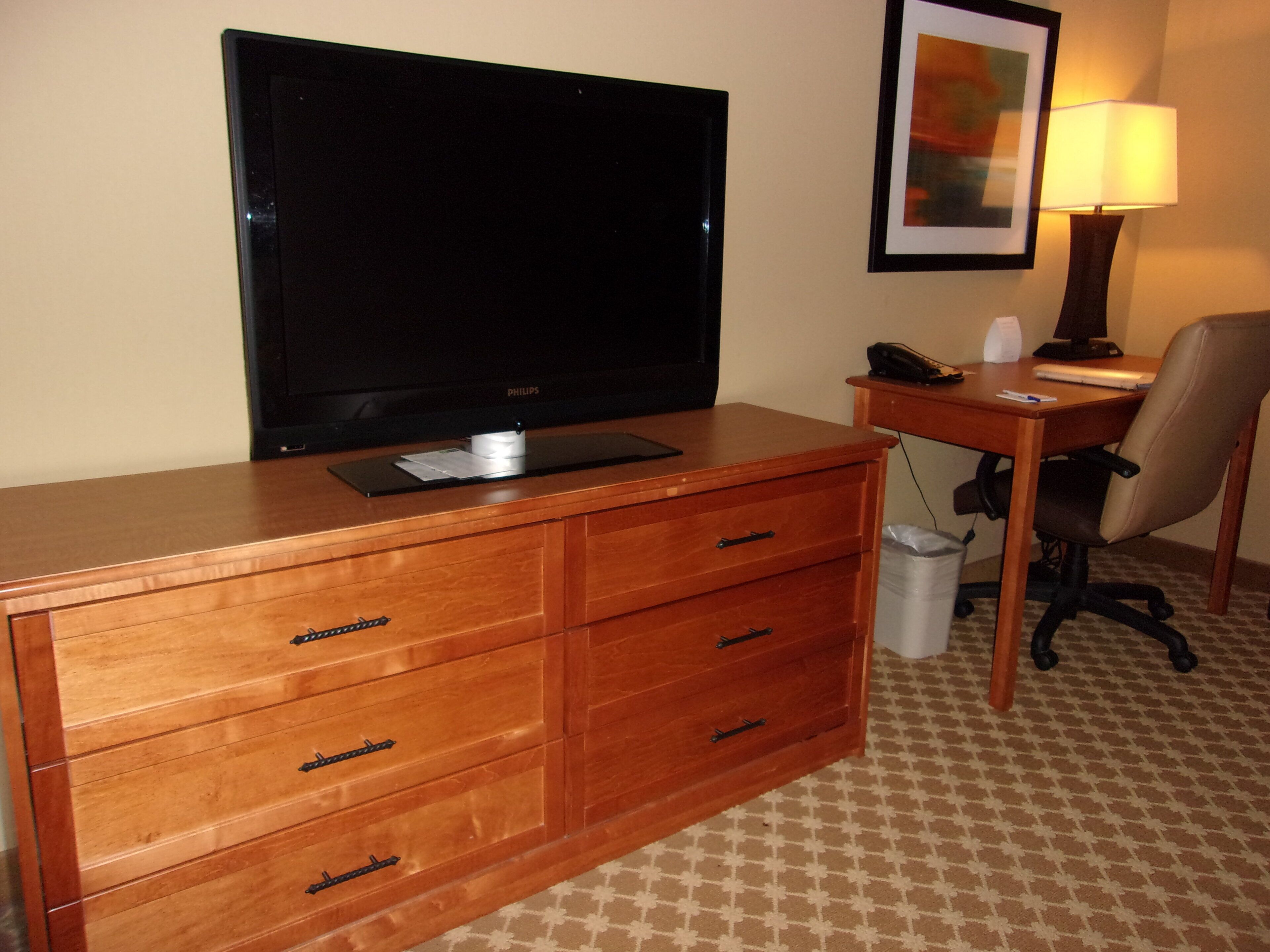 A hotel room with a wooden dresser, a flat-screen TV, a desk with a lamp, and a chair.