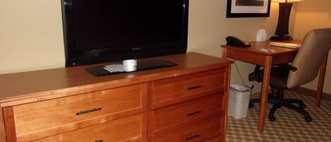A hotel room with a wooden dresser, a flat-screen TV, a desk with a lamp, and a chair.