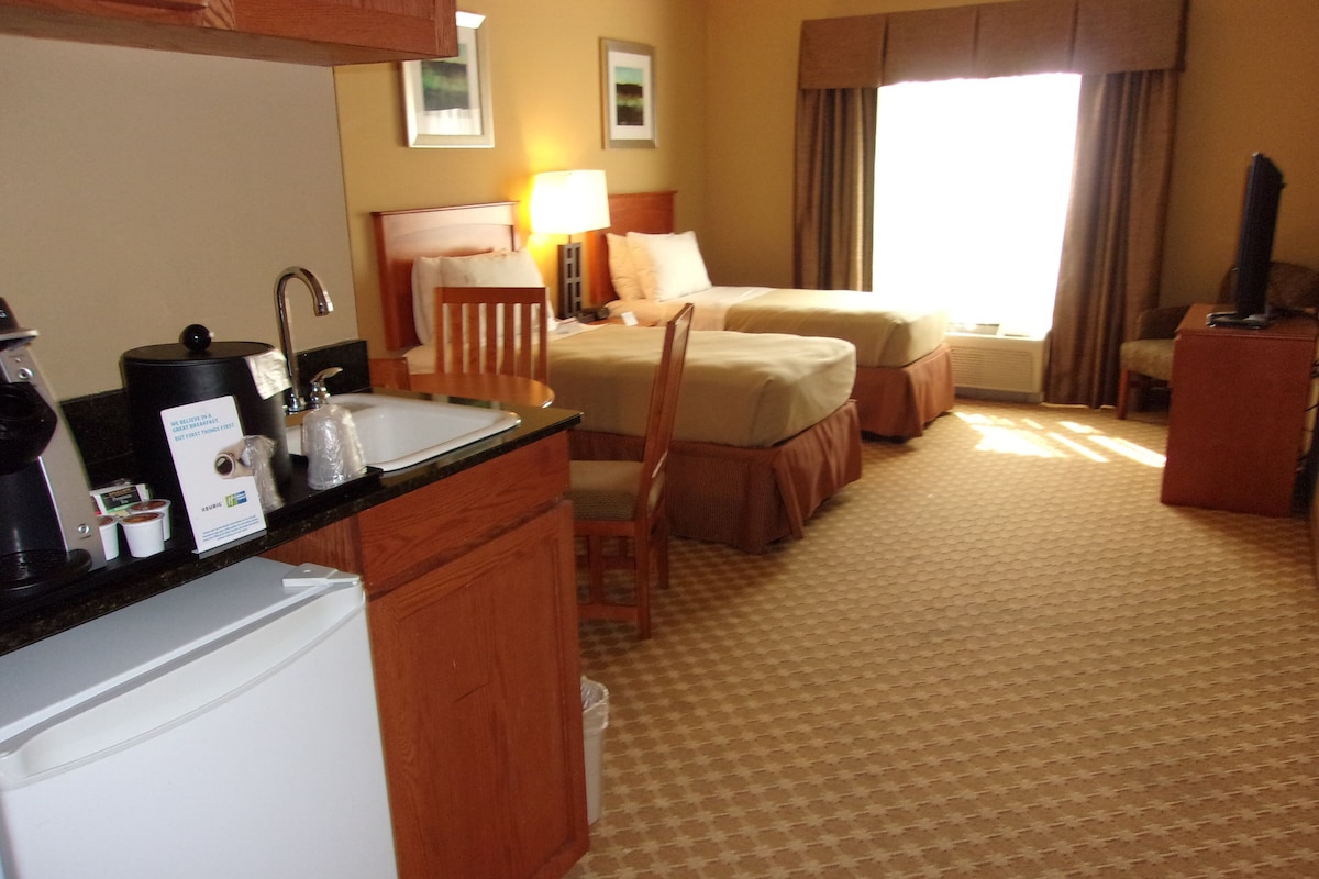 A hotel room with a kitchenette, a bed, a chair, a desk, and a television.