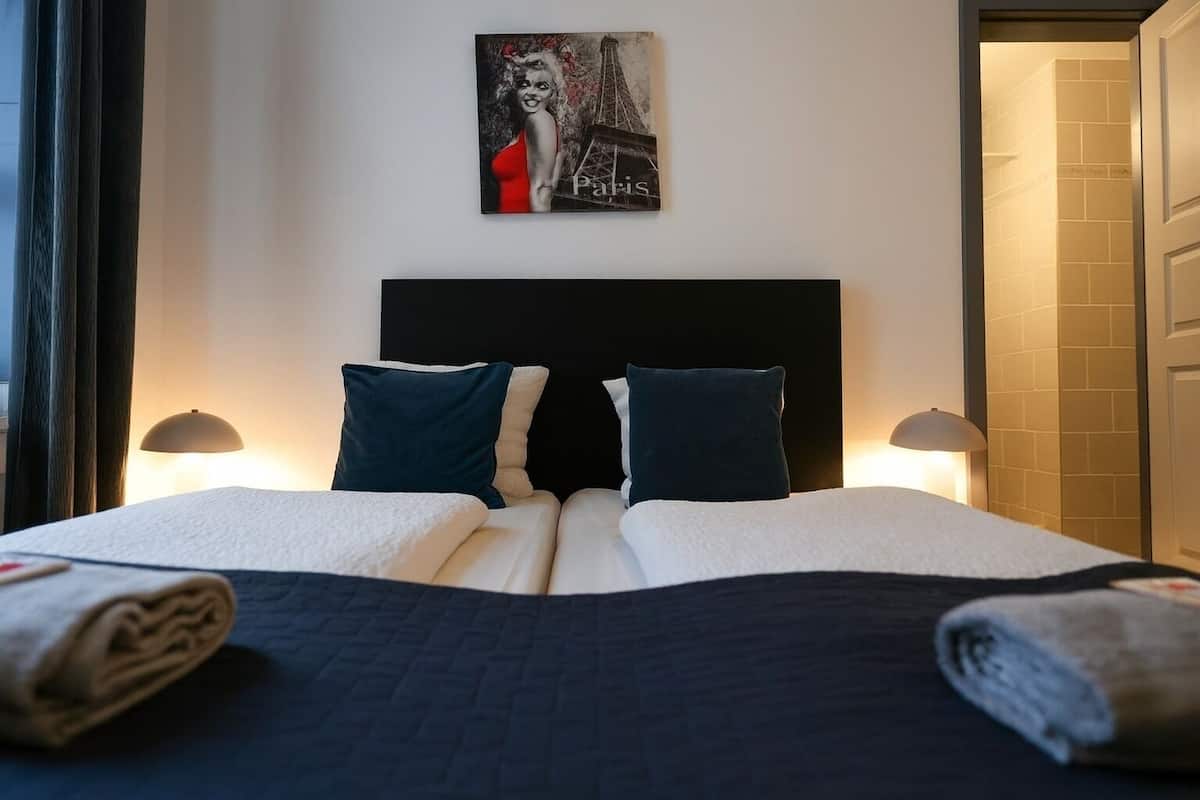 A neatly made bed with two pillows, a headboard, and a wall-mounted picture of a woman in a red dress with the word "Paris" below it.