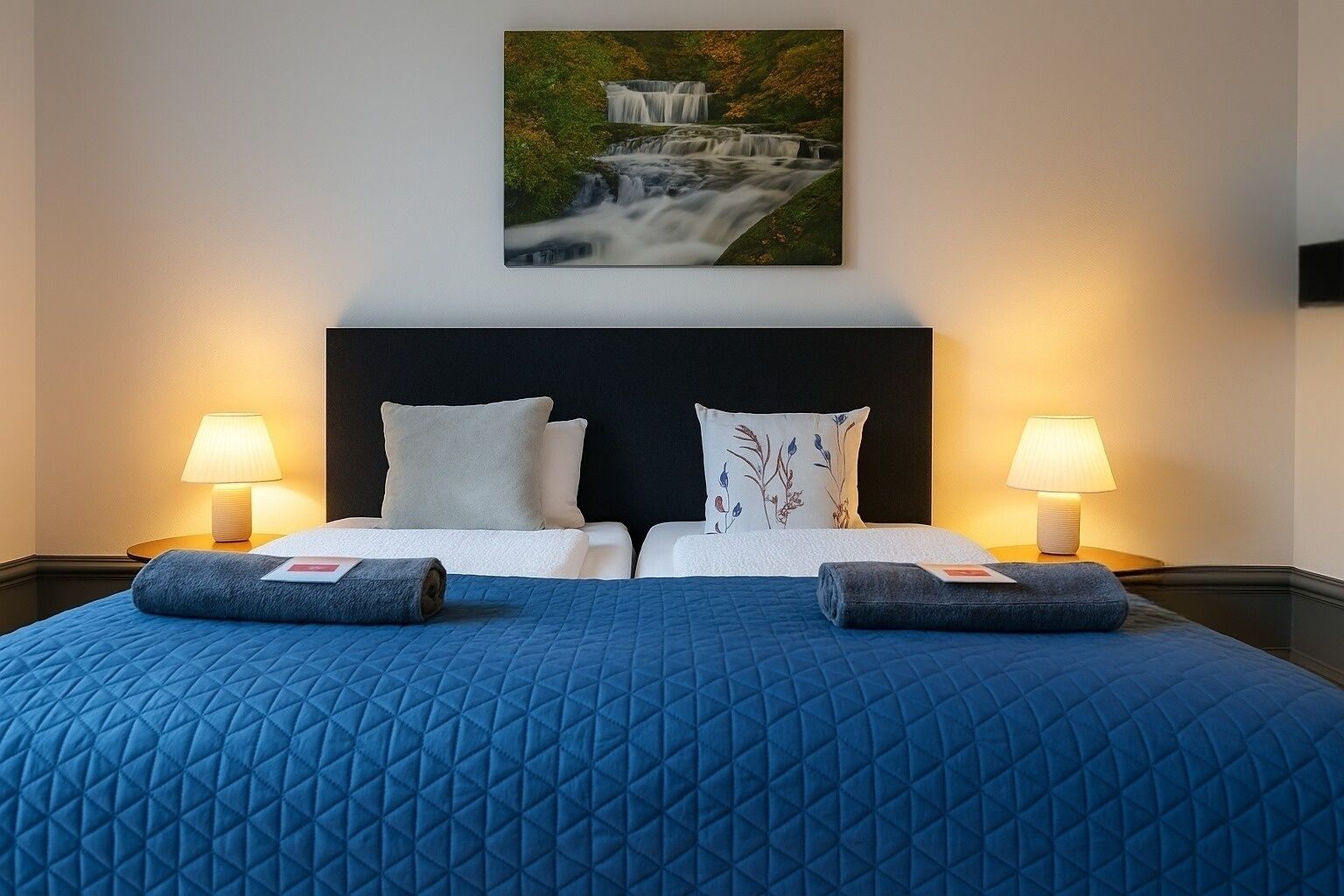 A double bed with a blue quilted bedspread, two pillows, and two bedside lamps.