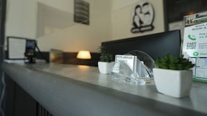A hotel reception desk with a clear desk area, a small potted plant, a sign for the hotel amager, and a digital screen displaying hotel information. - Hotel Amager (Copenhagen)