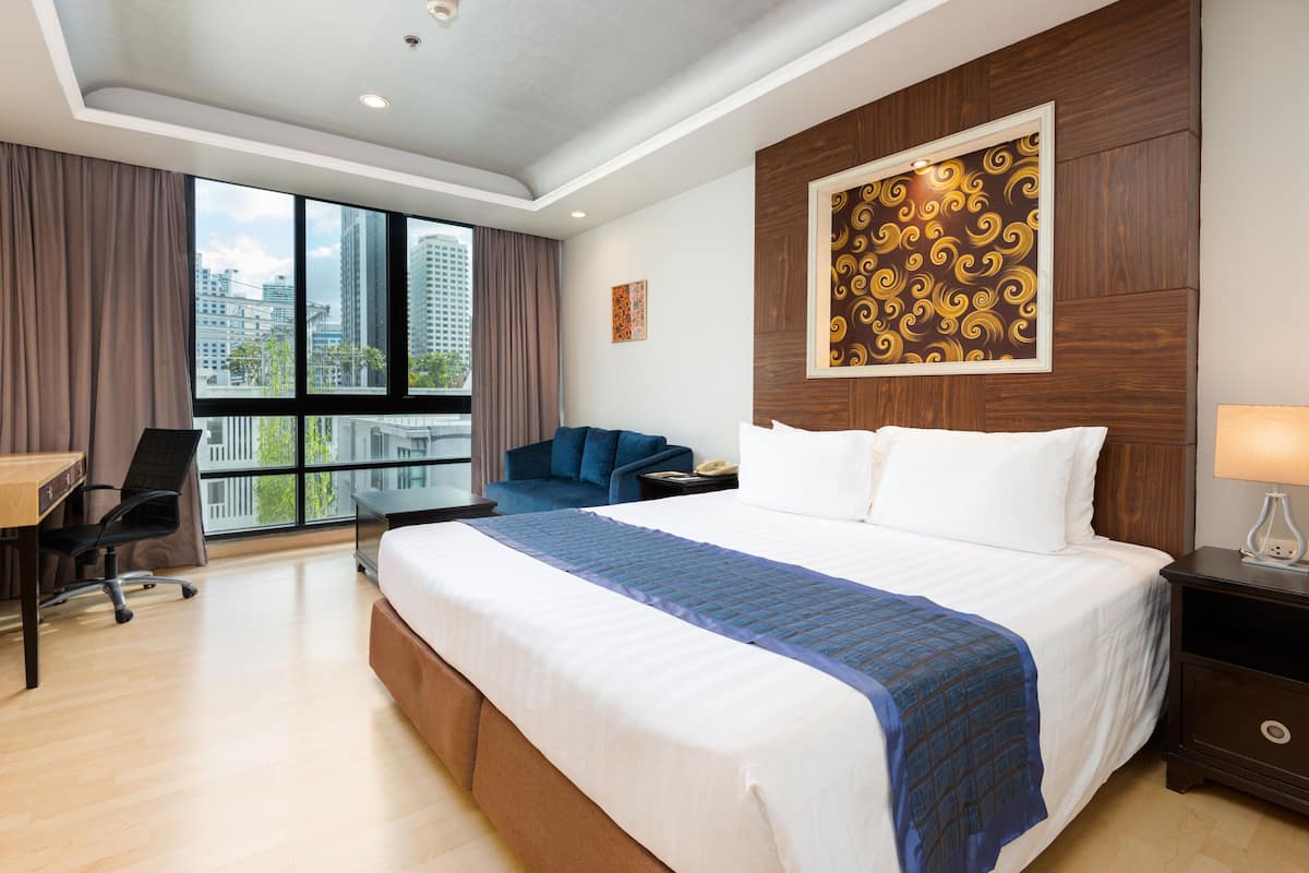 A hotel room with a large bed, a desk, a chair, and a view of the city through large windows.