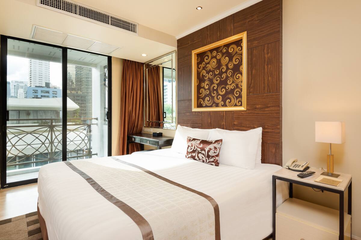 A hotel room with a large bed, a desk with a telephone, and a view of the city through a glass door.