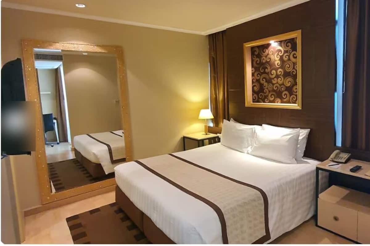 A hotel room with a large bed, a bedside table with a lamp, a mirror, and a wall-mounted artwork.