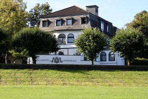 A large, historic building with a white facade and dark roof, surrounded by trees and a grassy area.