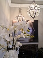 Una habitación con un cuadro grande de un barco en el agua, un jarrón con orquídeas blancas y dos lámparas colgantes.