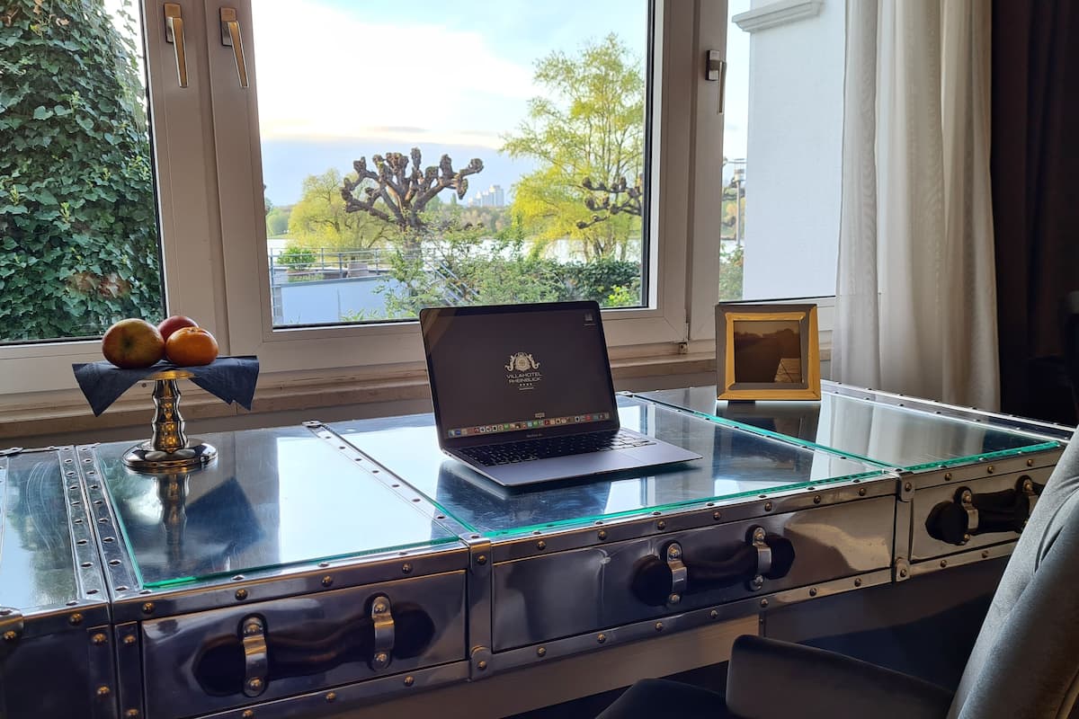 En bærbar computer på et glasbord med skuffer, en skål med frugt og udsigt til træer og bygninger.