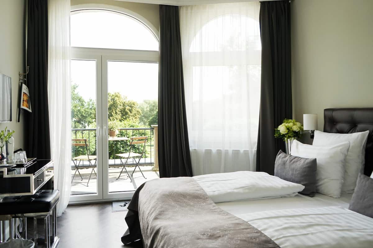 Et hotelværelse med seng, skrivebord, stol, balkon med bord og stole, samt vindue med gardiner.