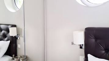 A modern hotel room with a large mirror, a round window, a bed with a dark headboard, and two side tables with lamps.