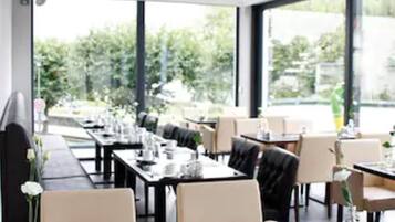 A modern dining area with tables set for service, large windows offering a view of greenery, and a clean, contemporary interior design.