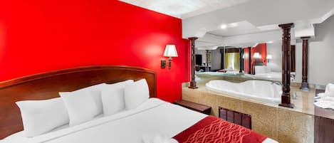 A hotel room with a large bed, a red accent wall, a bathroom with a bathtub, and a mirror.