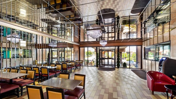 A spacious indoor area with multiple dining tables and chairs, a central open space, and a view through glass doors. - Rodeway Inn Capri (Little Ferry)