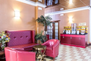 A lobby with a red sofa, a red armchair, a small round table, a potted plant, and a counter with a sign indicating guest rooms.