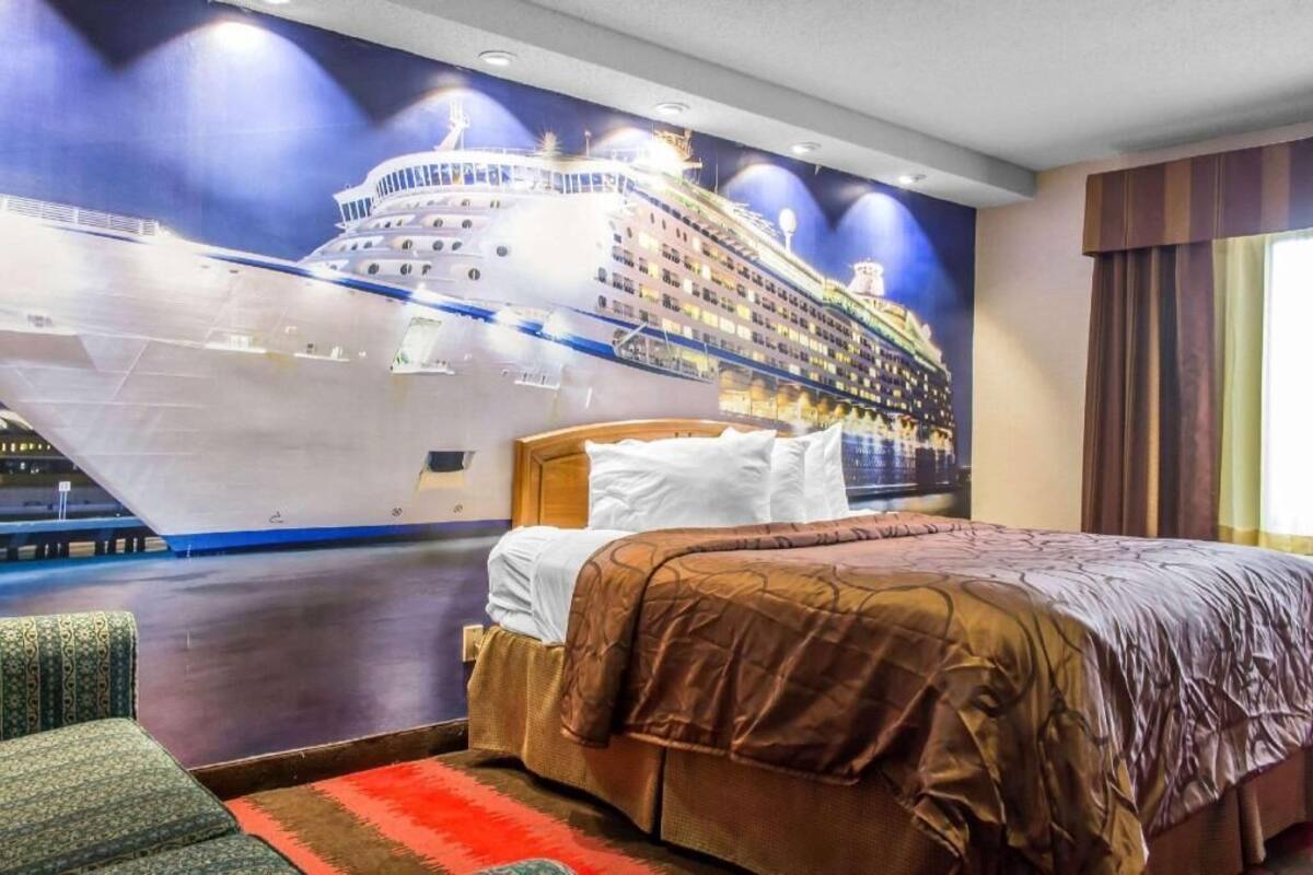 A hotel room with a large bed, a headboard, a wall mural of a cruise ship, and a window with curtains.