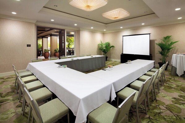 Un salón de conferencias con una larga mesa, sillas, una pantalla de proyector y plantas en macetas.